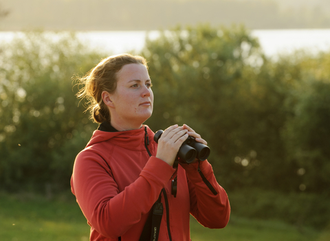 A woman standing in golden light of dawn or dusk appears to have just lowered binoculars from her face as she stares into the distance. Behind her is a row of trees and shore line of a lake.