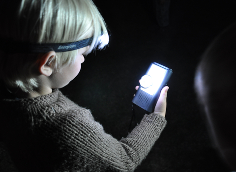 A young boy looks at a bat detector under the light of a headlamp.