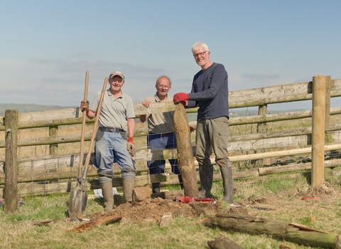 A group of three men aged in their 60s stand next to a fence in a countryside location. They hold tools and a fence post, which they are working to repair. The sky is blue and they all look happy.