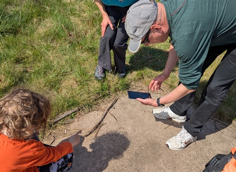 A woman stands watching as a man uses his phone to take a photo of something on the floor that a small child points at