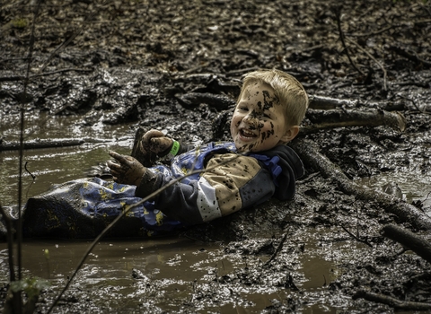A boy lies in mud/water and has it splattered over his face as he smiles