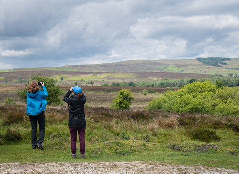 Two woman stand looking over moorland watching with binoculars and a camera. One wears a bright blue coat and black trousers while the other wears a blue cap and black coat. 