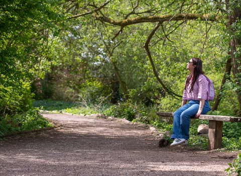 A woman sits on a wooden bench looking up towards the surrounding trees. She has long dark hair and wears blue jeans and a pink top.