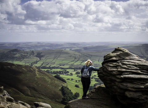 Girl with a rucksack on her back, holding onto a boulder looking out to a green landscape with rolling white clouds in the sky.