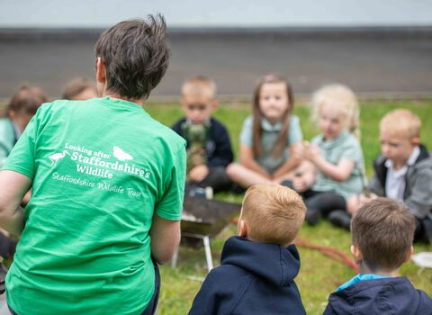 A woman sits at the head of a circle of school children. She wears a bright green tshirt that says Looking after Staffordshire's Wildlife on it