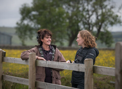 Two woman stand by a wooden fence in a field full of buttercups with trees in the distance. One has dark hair which they wear up, while the other has red/ginger hair which is worn down.