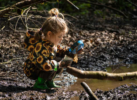 A young girl crouches next to a muddy puddle and pours water/mud from a saucer into a pot