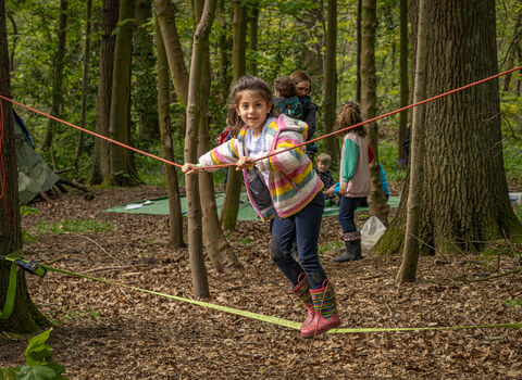 A young girl with dark brown hair stands balancing on a rope between two trees in a woodland setting