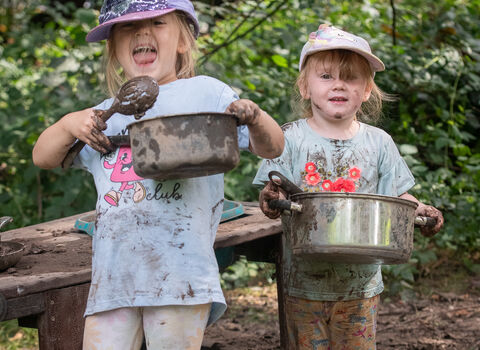 Two young girls smile as they hold up bots covered in mud as they play in a mud kitchen. Their clothing is covered in mud and they're in a an outdoor forest school setting.