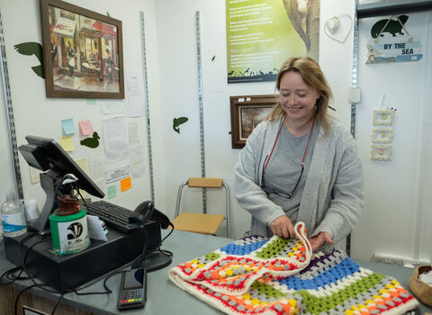 A woman at a till in a charity shop folding a colourful crochet blanket.