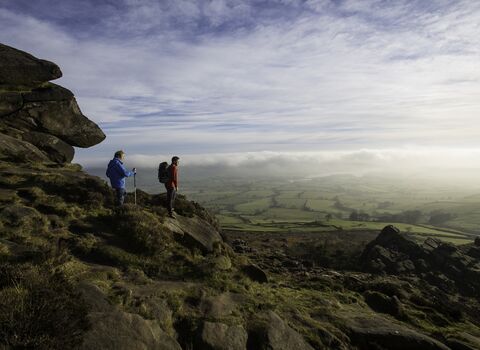 Two men stand overlooking a vast landscape from high on a hill with rocks behind them