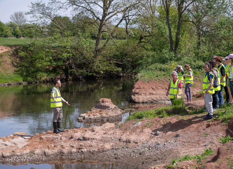 A group of people in high vis on a nature reserve, next to a river where a man is standing speaking to them.